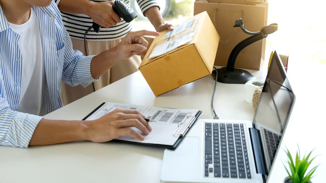 Person scans a package with a barcode scanner next to a laptop on a desk. Another individual points at a clipboard, indicating package management.