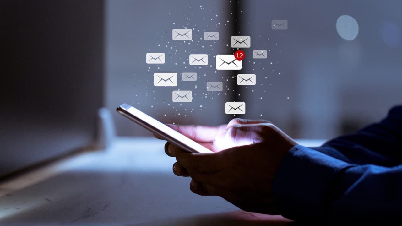 A person using a smartphone in a dimly lit room with digital email icons hovering above, including a red notification showing 12 new messages.
