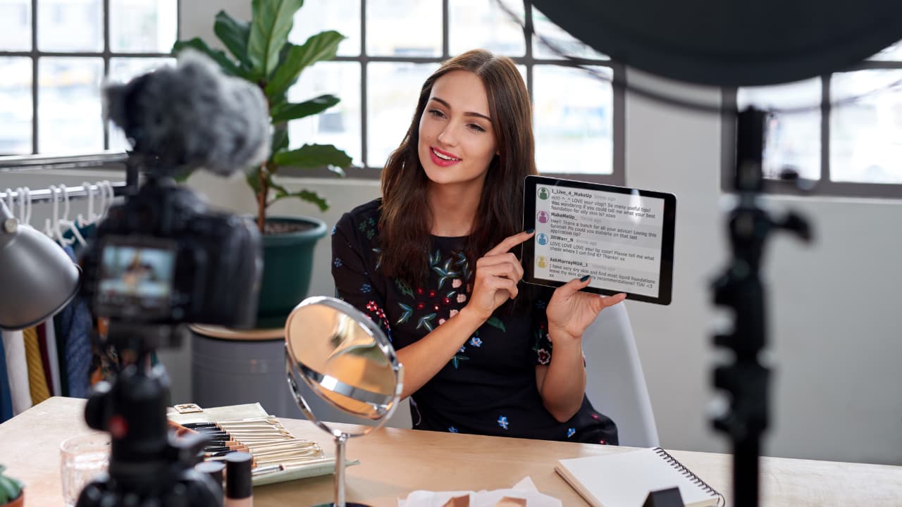A woman smiles at the camera while holding a tablet displaying text. She's in a bright room with a camera, plants, and makeup items, suggesting a vlog setting.