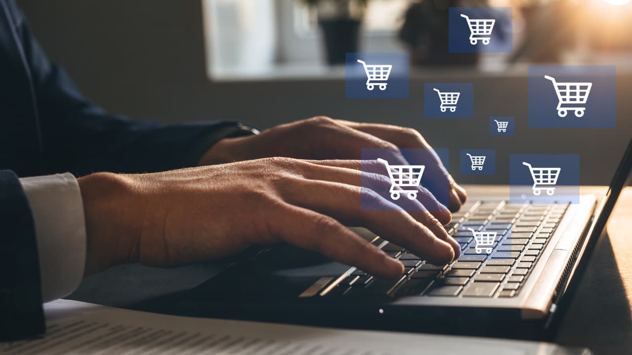 Hands typing on a laptop keyboard with digital shopping cart icons floating above, suggesting online shopping. The scene conveys a modern, tech-savvy vibe.