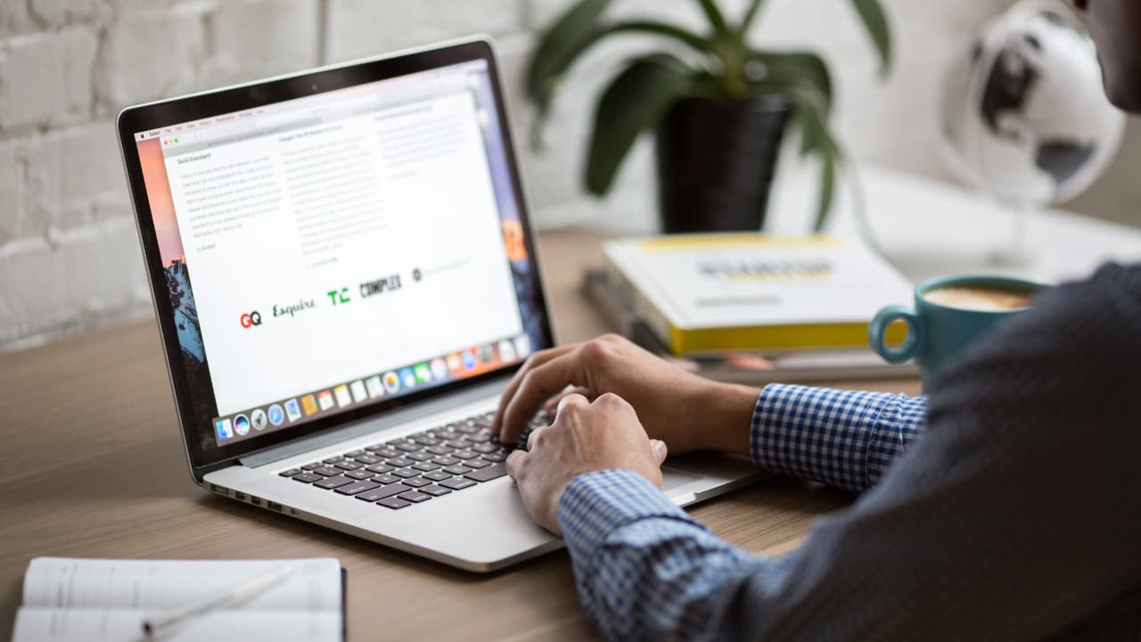 A person types on a laptop at a wooden desk, surrounded by books and a plant, conveying a focused and productive work environment.