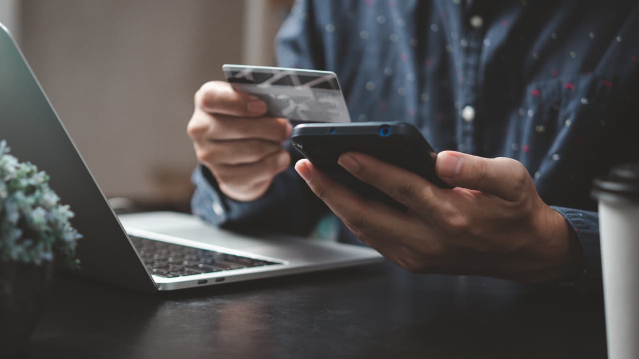 A person holds a credit card and smartphone at a desk with a laptop nearby, suggesting online shopping or mobile banking. The tone is focused and modern.