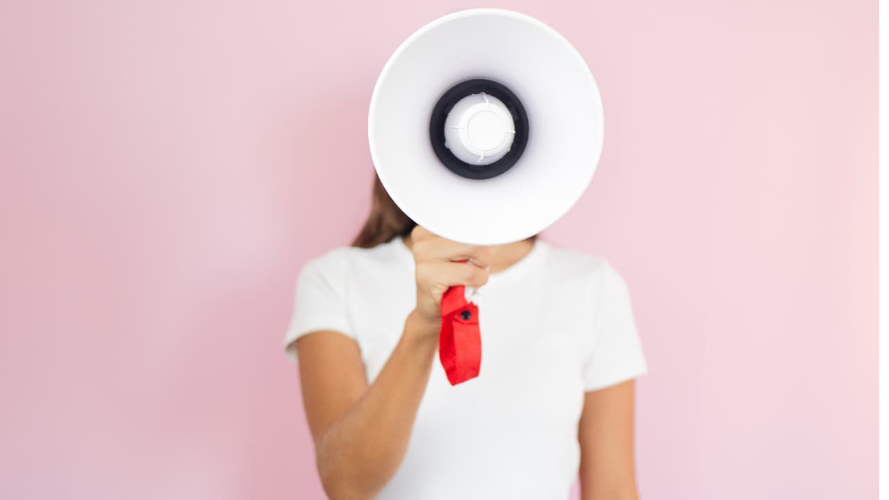 Person holding a megaphone in front of their face against a pink background, wears a white shirt. The mood is proactive and assertive.