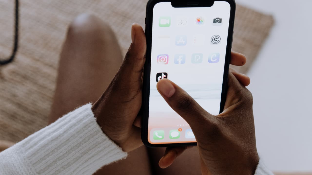 Close-up of hands holding a smartphone. The phone screen displays social media apps, including TikTok. The mood is casual and focused.