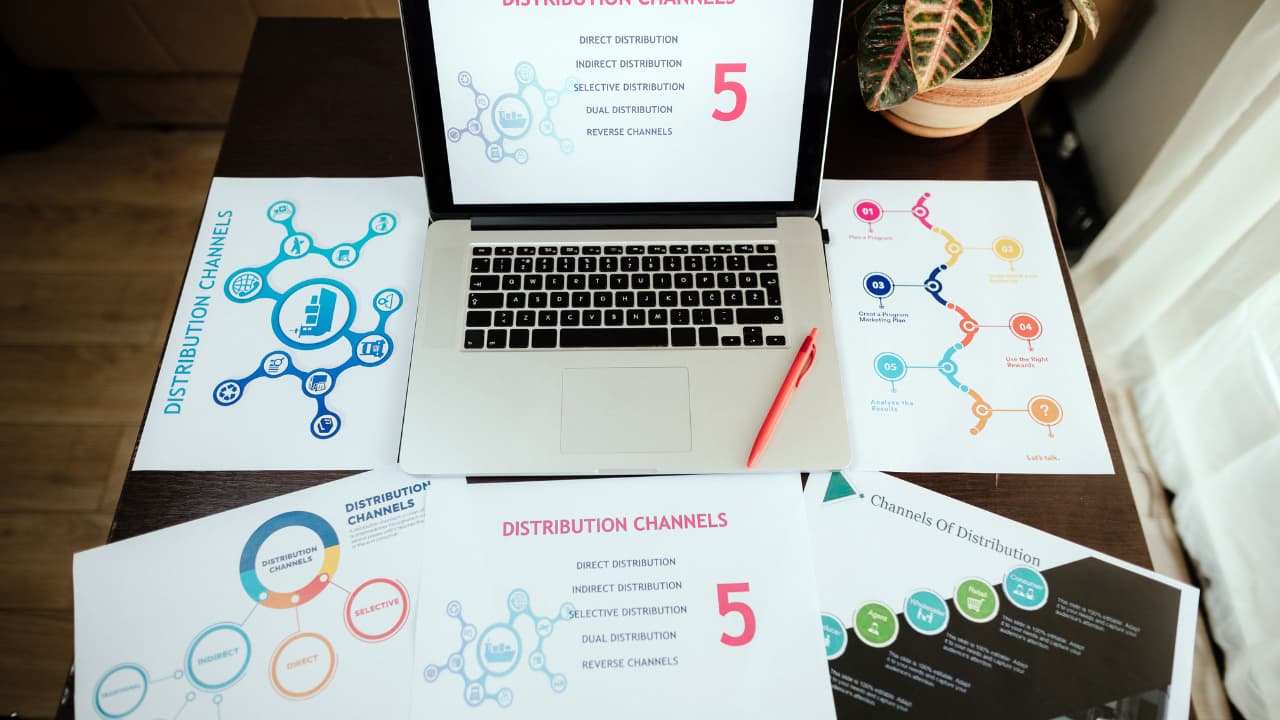 Laptop on a desk displaying "Distribution Channels." Surrounding papers show colorful distribution charts. A red pen lies beside the laptop, creating a business-focused atmosphere.