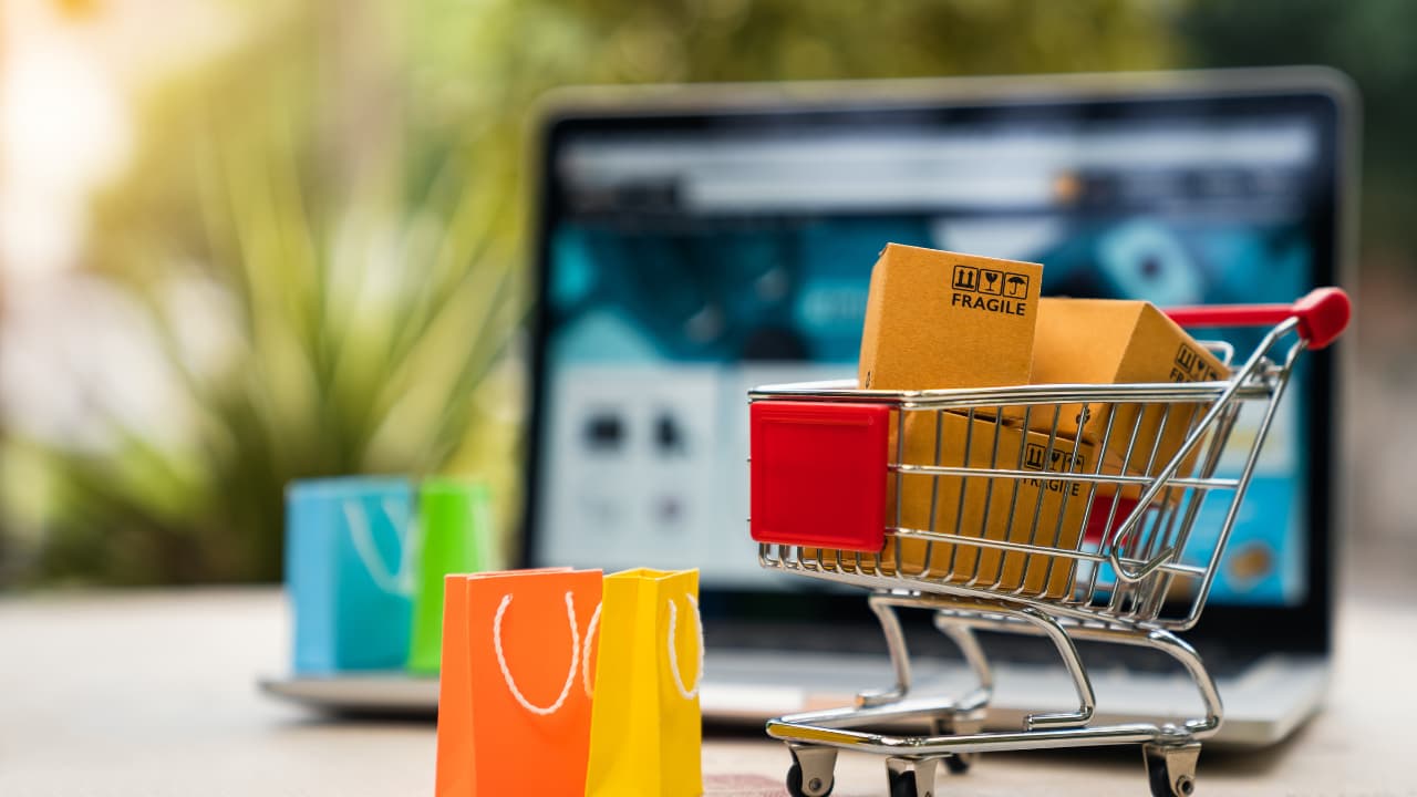 Mini shopping cart with small parcels marked "fragile" on a desk. Colorful gift bags nearby, laptop with blurred screen in background, suggesting online shopping.
