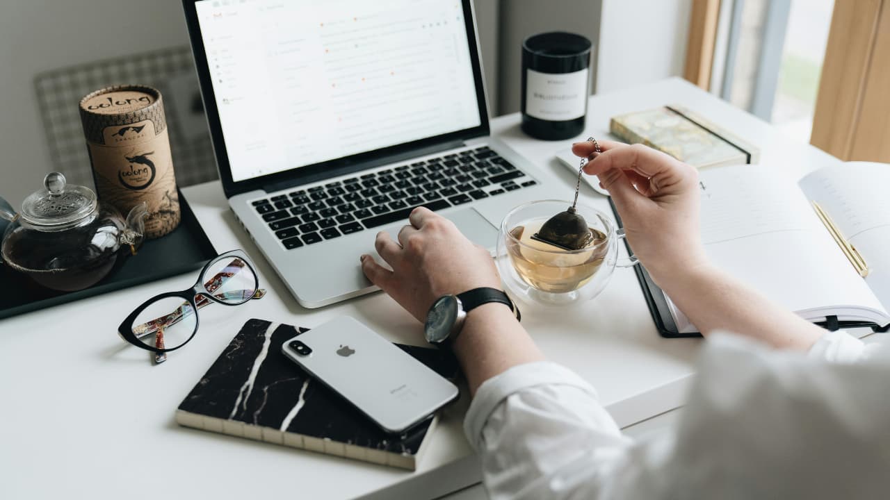 A person stirs tea at a stylish desk with a laptop, phone, glasses, and a notebook. The scene feels calm and organized, ideal for focused work.
