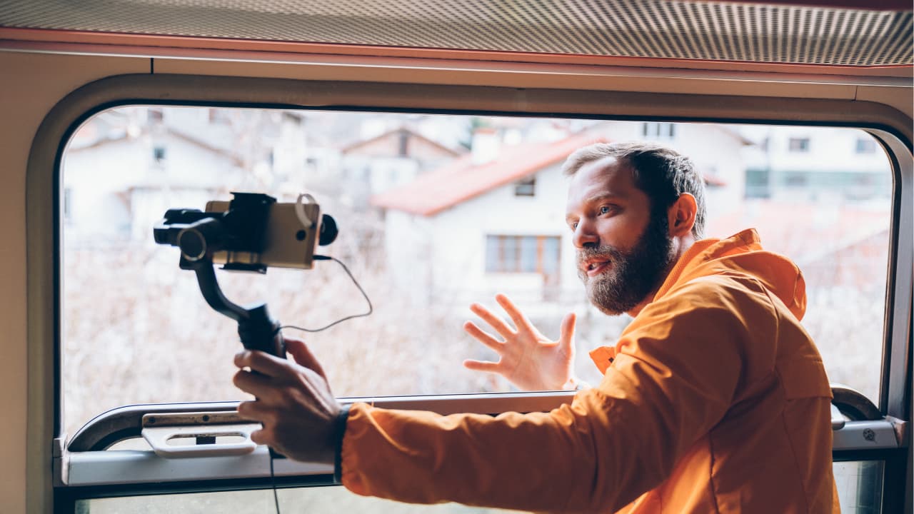 A man in an orange jacket enthusiastically gestures while filming with a camera on a stabilizer through a train window. Blurred houses are visible outside.