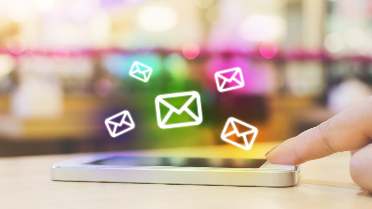 A smartphone on a table with a finger touching the screen. Colorful, glowing email icons float above, suggesting digital communication and connectivity.