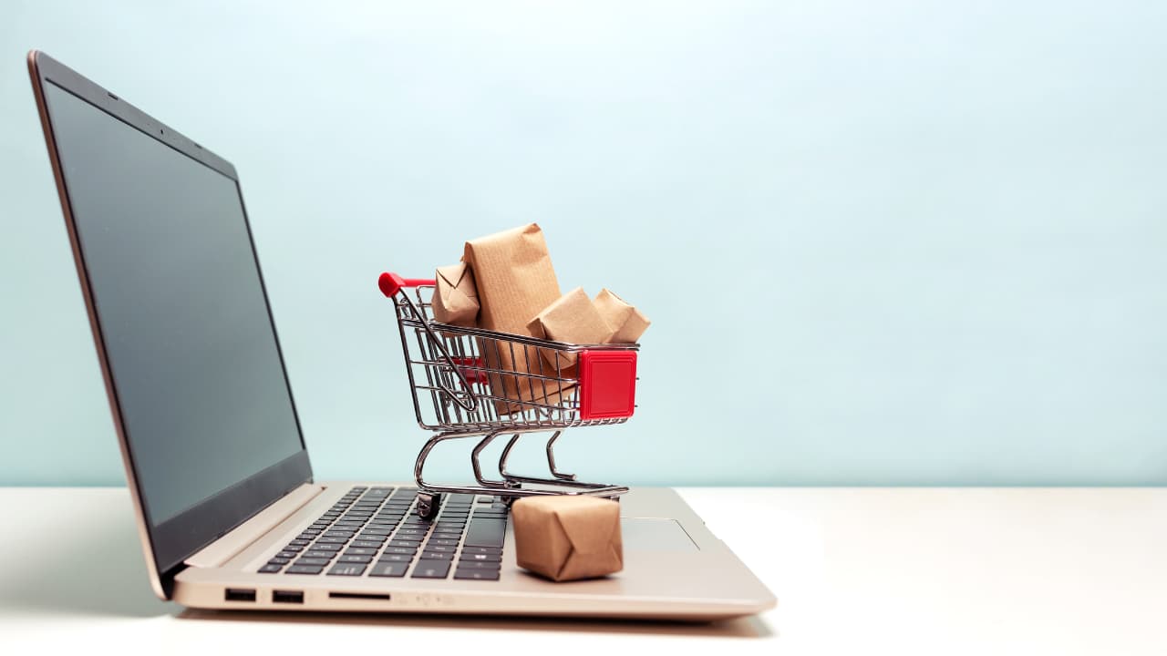 An open laptop with a miniature shopping cart on its keyboard, filled with small brown packages. The background is a soft blue, conveying online shopping.