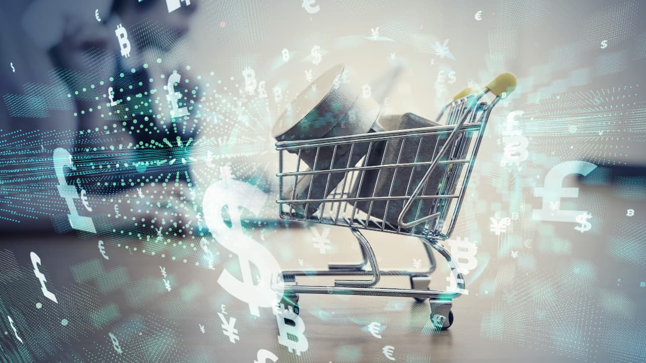 A small shopping cart filled with stacked coins is surrounded by floating currency symbols like dollar and euro, symbolizing global commerce and finance.