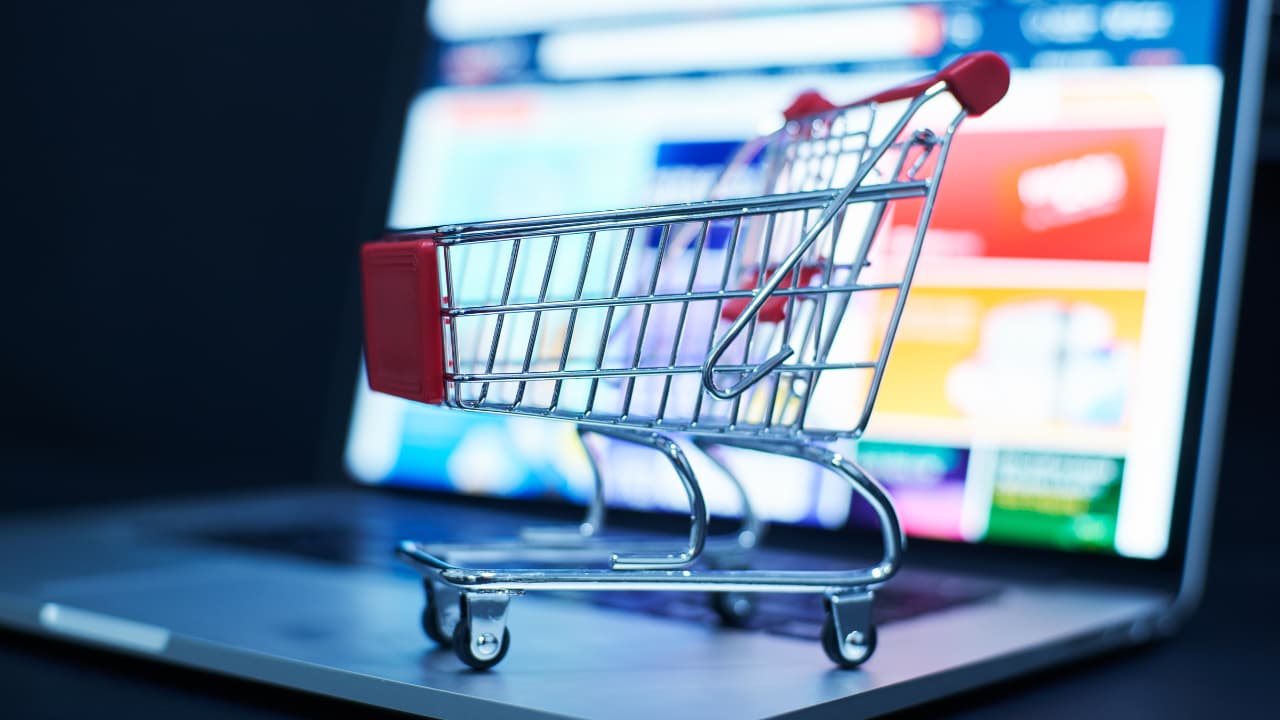 A small shopping cart sits on a laptop keyboard, with a blurred screen displaying colorful shopping website graphics. The scene conveys online shopping.