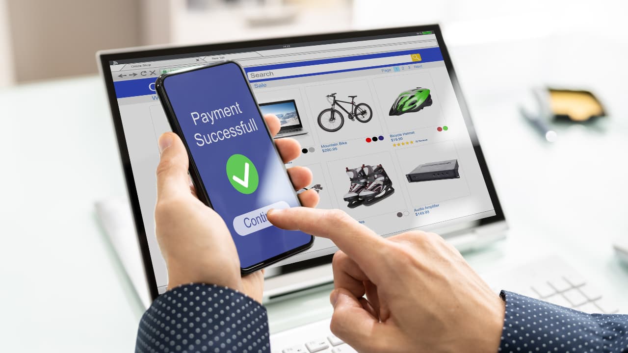 A person holds a smartphone showing "Payment Successful" with a checkmark. Laptop in background displays an online shopping page with various products.