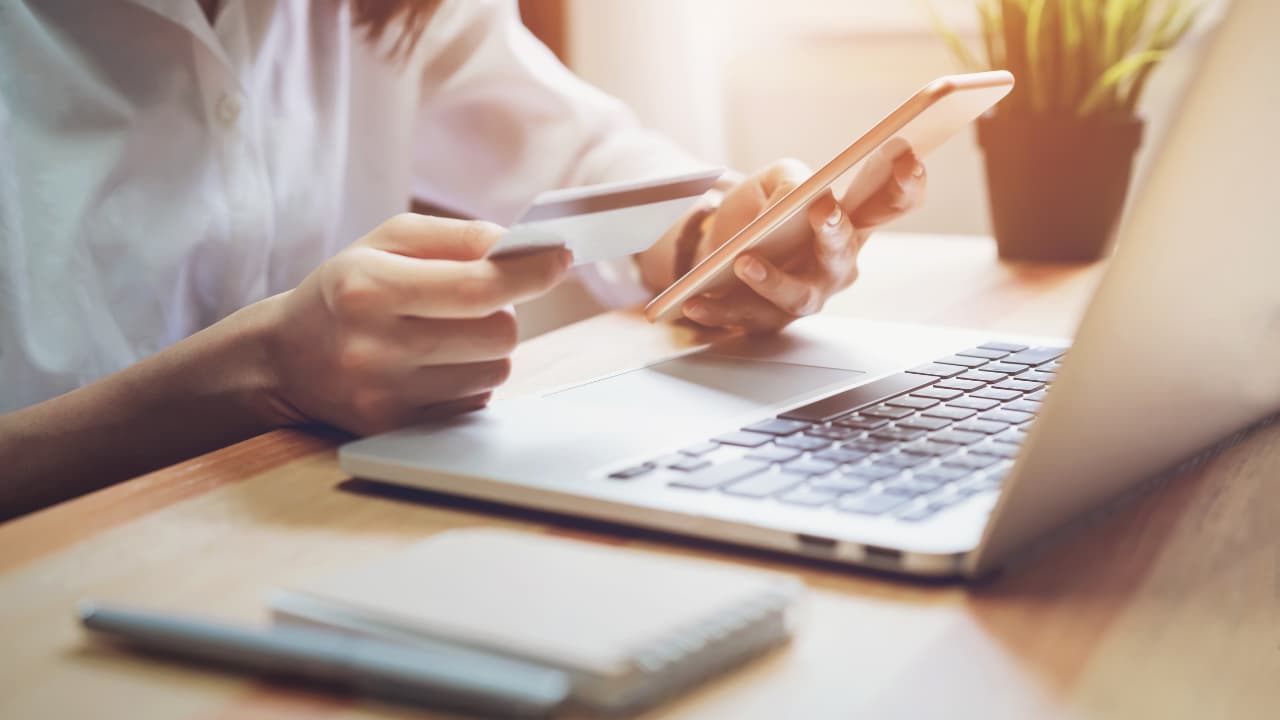 A person wearing a white shirt holds a credit card and a smartphone near an open laptop on a wooden desk. A plant and notebook are in the background, suggesting productivity.