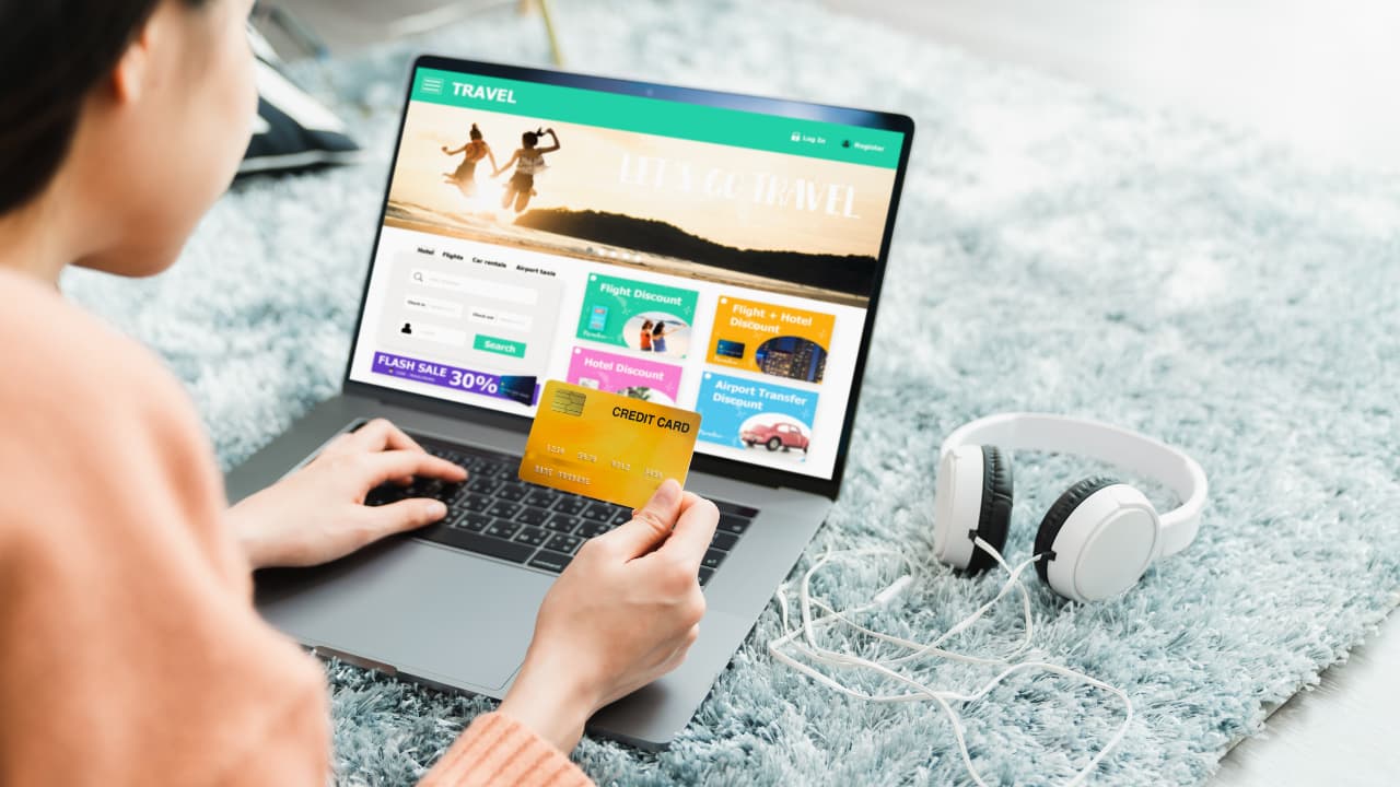 A person holds a credit card while shopping for travel deals on a laptop. The screen displays "Let's Go Travel," colorful offers, and images. Headphones lie nearby on a plush rug.