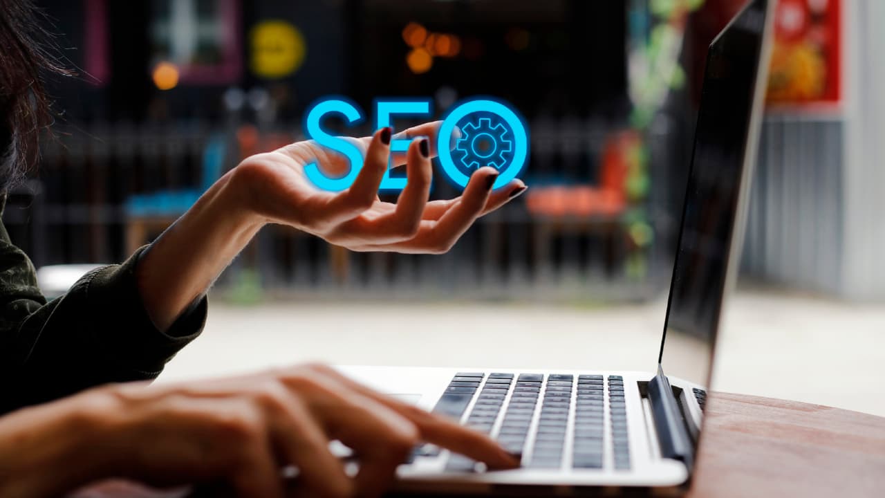 Person using a laptop with a floating "SEO" graphic above their hand. The image conveys technology and digital marketing concepts. Background is blurred.