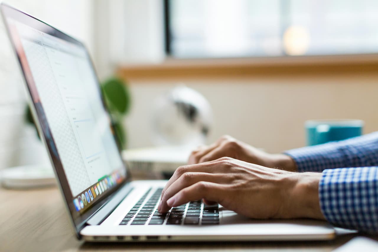 Hands typing on a laptop keyboard in a bright workspace. The screen displays a document. A blue coffee mug and green plant in the background convey focus.