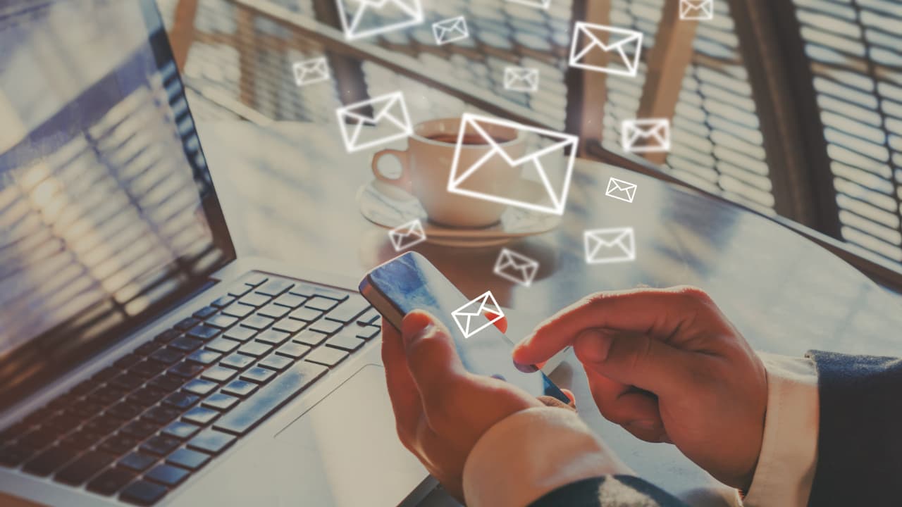 A person holds a smartphone, interacting with floating email icons, next to a laptop on a sunlit table, suggesting active communication.