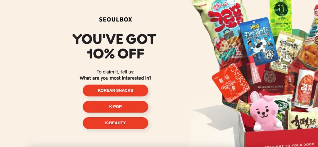 SeoulBox promotion offering 10% off. Left text asks preference: Korean snacks, K-pop, or K-beauty. Right features a box filled with Korean snacks, colorful wrappers, and a pink bunny toy.