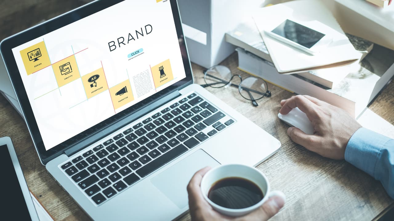 A person works at a wooden desk, viewing a laptop screen displaying branding concepts. They hold a coffee cup, conveying focus and productivity.