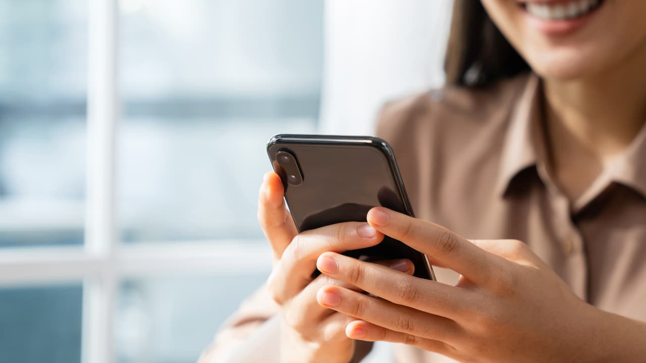 A woman smiles as she uses a smartphone indoors. Her hands hold the phone, displaying engagement. Soft lighting and a blurred background create a warm ambiance.