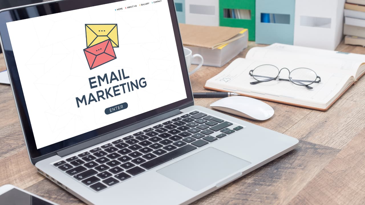 A laptop on a wooden desk displays a webpage titled "Email Marketing" with colored envelopes. Surrounding items include a notebook, glasses, and office supplies.