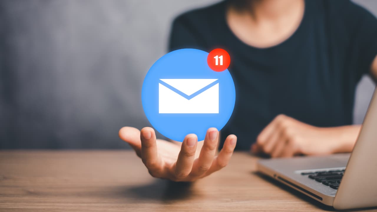 A person in a dark shirt is sitting at a wooden desk with a laptop. They hold a floating email icon showing 11 unread messages, conveying a sense of digital connectivity.