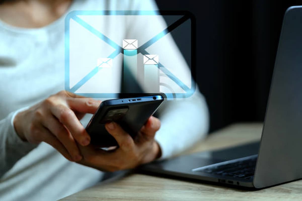 A person in a white sweater uses a smartphone near a laptop. A digital envelope with icons of flying emails appears, suggesting communication technology.