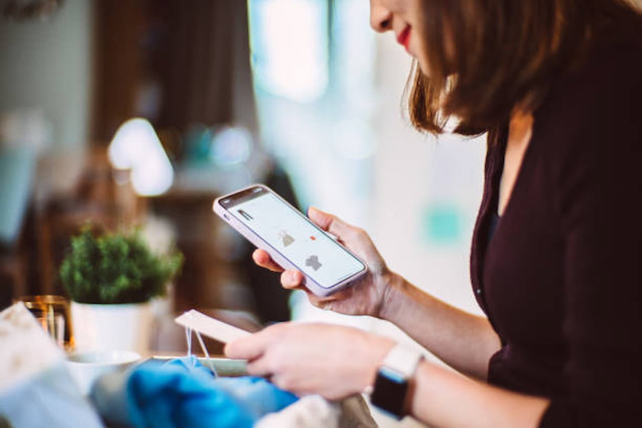 A woman in a dark sweater is shopping online with a smartphone, holding a credit card. The setting is cozy, with a blurred background and soft lighting.