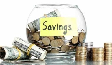 A glass jar labeled "Savings" is filled with coins, surrounded by stacks of coins and rolled dollar bills. The image conveys financial planning and security.
