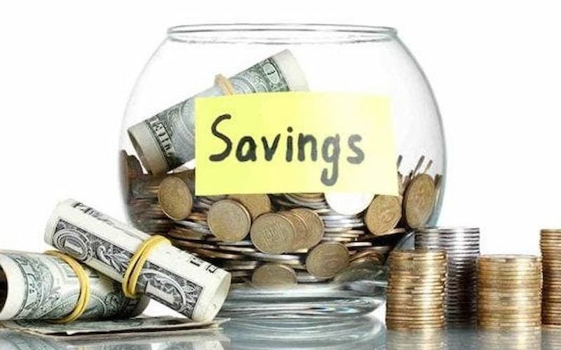 A glass jar labeled "Savings" is filled with coins, surrounded by stacks of coins and rolled dollar bills. The image conveys financial planning and security.